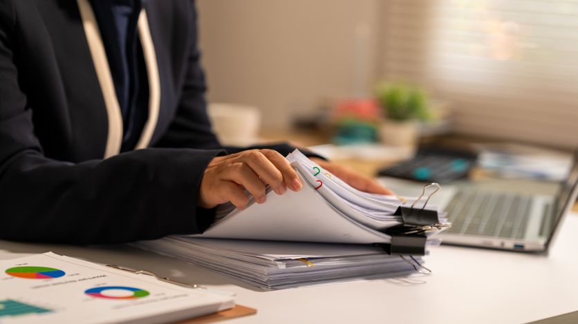 Businesswoman reviewing a stack of documents at an office desk with charts, binder clips and laptop, showing paperwork, analysis and everyday administrative work. L’impact de l’intelligence artificielle sur l’emploi des jeunes diplômés